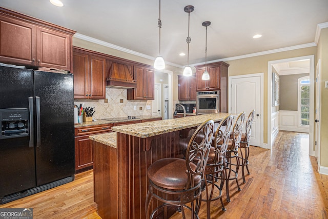 kitchen with light hardwood / wood-style floors, ornamental molding, black appliances, and an island with sink