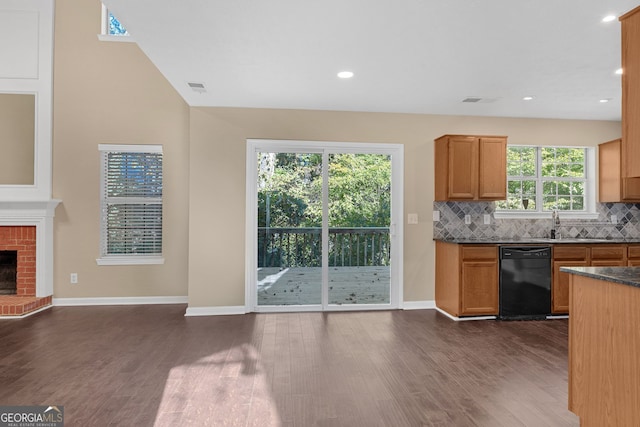 kitchen with black dishwasher, dark hardwood / wood-style flooring, a brick fireplace, dark stone countertops, and decorative backsplash