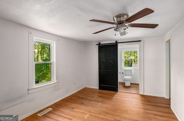 unfurnished bedroom featuring ceiling fan, a textured ceiling, a barn door, connected bathroom, and light hardwood / wood-style floors