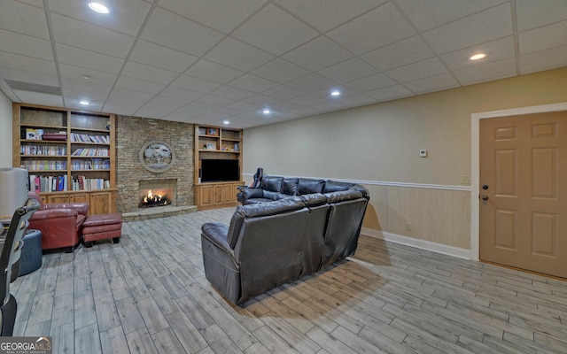 living room featuring a drop ceiling, a stone fireplace, built in shelves, and light wood-type flooring