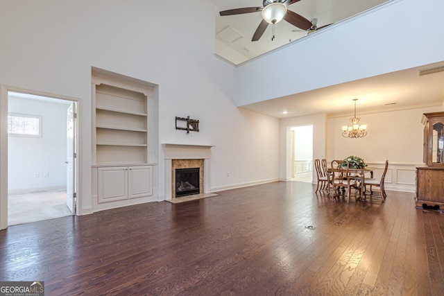 living room with ceiling fan with notable chandelier, a towering ceiling, a tiled fireplace, dark hardwood / wood-style flooring, and built in shelves