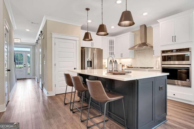 kitchen featuring wall chimney range hood, an island with sink, stainless steel appliances, hardwood / wood-style floors, and white cabinetry