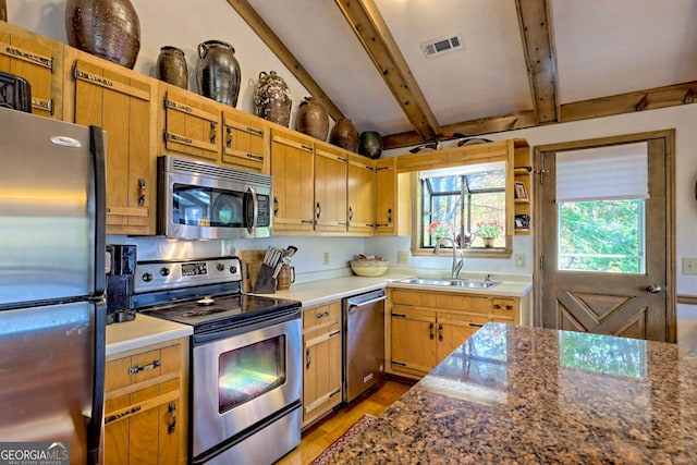 kitchen with beam ceiling, appliances with stainless steel finishes, light wood-type flooring, dark stone countertops, and sink