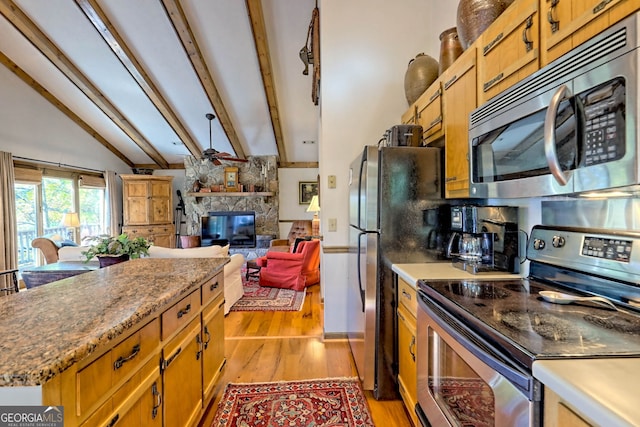 kitchen featuring a stone fireplace, vaulted ceiling with beams, light hardwood / wood-style flooring, appliances with stainless steel finishes, and ceiling fan