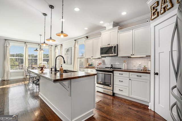 kitchen with hanging light fixtures, appliances with stainless steel finishes, white cabinetry, a kitchen island with sink, and wooden counters