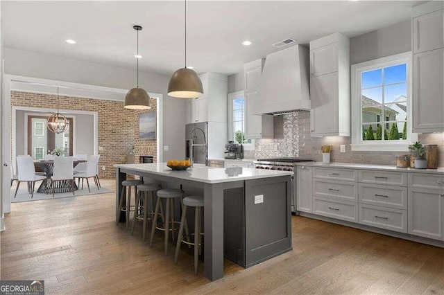 kitchen with custom exhaust hood, hanging light fixtures, a kitchen island with sink, light wood-type flooring, and stainless steel appliances