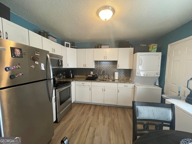 kitchen with appliances with stainless steel finishes, sink, white cabinets, stacked washing maching and dryer, and light hardwood / wood-style flooring