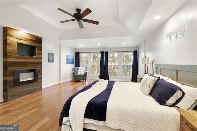 bedroom featuring wood finished floors, ceiling fan, a large fireplace, a raised ceiling, and recessed lighting