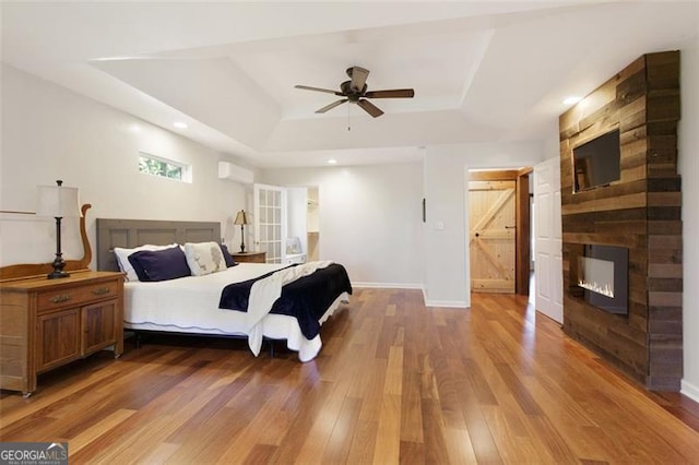 bedroom featuring a wall mounted air conditioner, a large fireplace, a raised ceiling, ceiling fan, and wood-type flooring