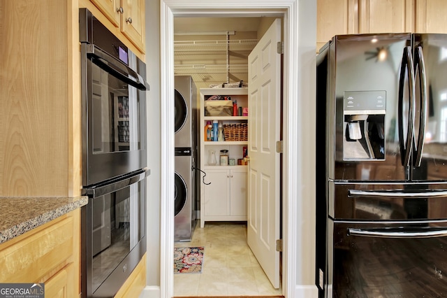 kitchen with light brown cabinets, stacked washer and dryer, black double oven, light tile patterned floors, and stainless steel fridge with ice dispenser