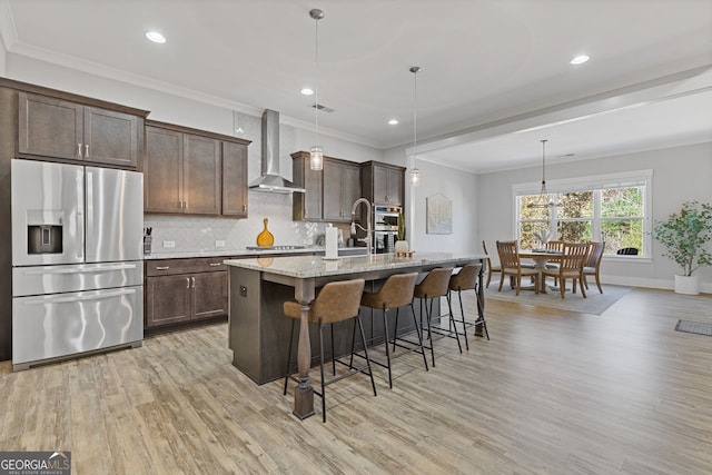 kitchen featuring backsplash, appliances with stainless steel finishes, wall chimney range hood, dark brown cabinets, and an island with sink