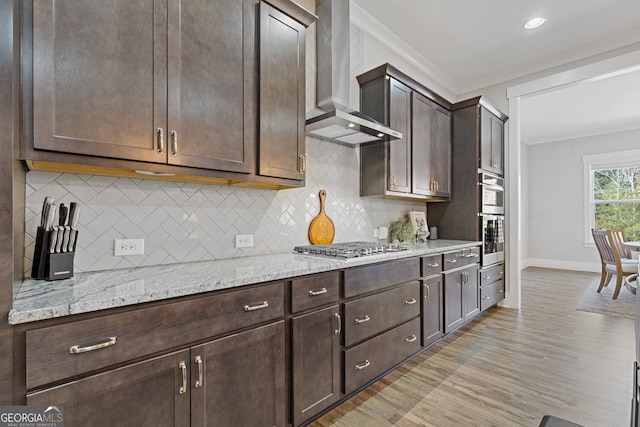 kitchen featuring crown molding, light wood finished floors, stainless steel appliances, wall chimney range hood, and dark brown cabinets