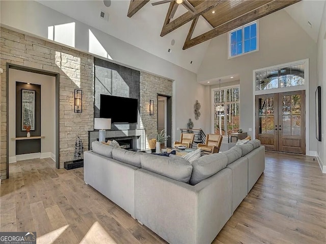 living room with beam ceiling, high vaulted ceiling, light hardwood / wood-style flooring, a stone fireplace, and french doors