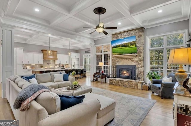 living room featuring ceiling fan, light wood-type flooring, beamed ceiling, a stone fireplace, and coffered ceiling