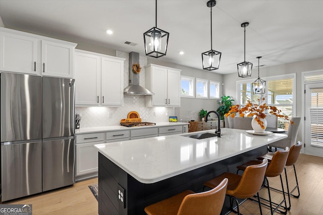 kitchen featuring pendant lighting, white cabinetry, wall chimney range hood, stainless steel appliances, and an island with sink