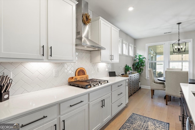 kitchen with pendant lighting, white cabinets, wall chimney exhaust hood, stainless steel gas cooktop, and light hardwood / wood-style flooring