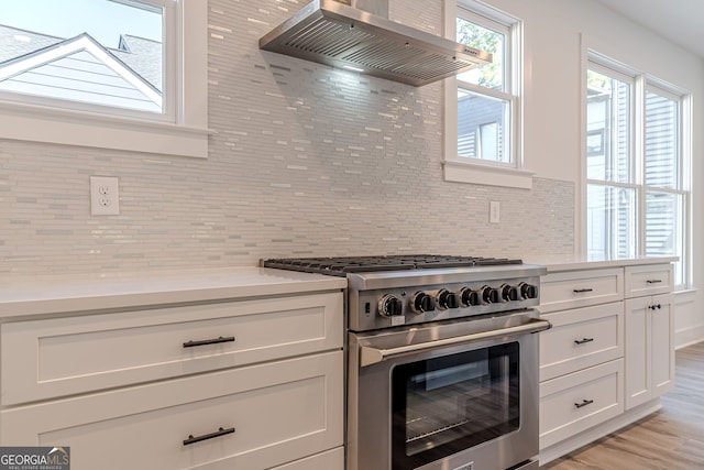 kitchen with wall chimney exhaust hood, high end range, white cabinets, light hardwood / wood-style floors, and tasteful backsplash