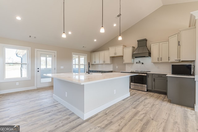 kitchen with a center island with sink, stainless steel stove, light wood-type flooring, custom range hood, and decorative light fixtures