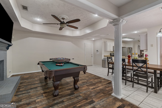 game room featuring ceiling fan, a textured ceiling, light wood-type flooring, a brick fireplace, and ornate columns