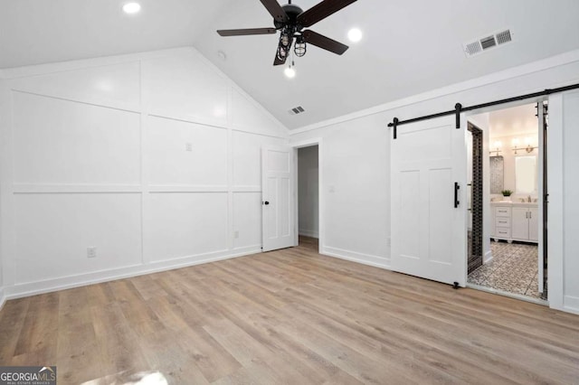 unfurnished bedroom featuring lofted ceiling, ceiling fan, a barn door, light wood-type flooring, and ensuite bath