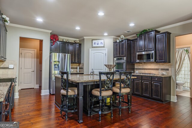 kitchen with dark hardwood / wood-style flooring, appliances with stainless steel finishes, an island with sink, crown molding, and light stone counters