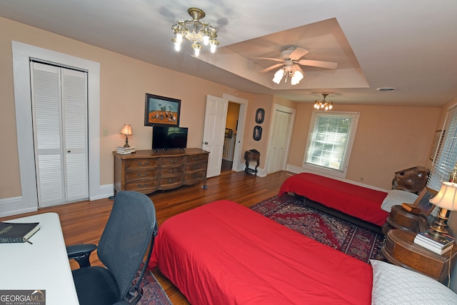 bedroom with dark wood-type flooring, a raised ceiling, multiple closets, and ceiling fan with notable chandelier