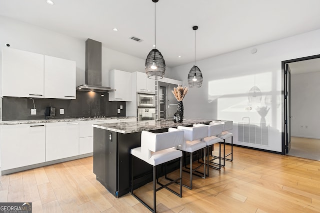 kitchen featuring wall chimney exhaust hood, stainless steel appliances, a center island with sink, decorative light fixtures, and white cabinetry