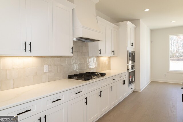 kitchen featuring custom exhaust hood, backsplash, appliances with stainless steel finishes, light hardwood / wood-style floors, and white cabinetry