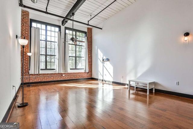 unfurnished living room with ceiling fan, beam ceiling, a high ceiling, wood-type flooring, and brick wall