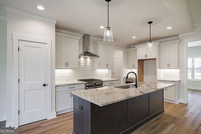 kitchen with sink, high end stainless steel range oven, wall chimney range hood, hanging light fixtures, and an island with sink