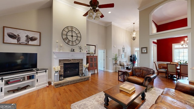 living area with a towering ceiling, wood finished floors, crown molding, a stone fireplace, and ceiling fan with notable chandelier