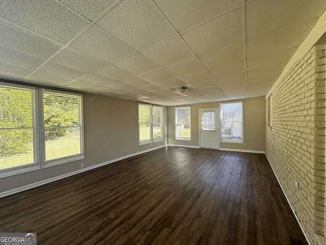 unfurnished living room featuring dark wood-style flooring, baseboards, a drop ceiling, and brick wall