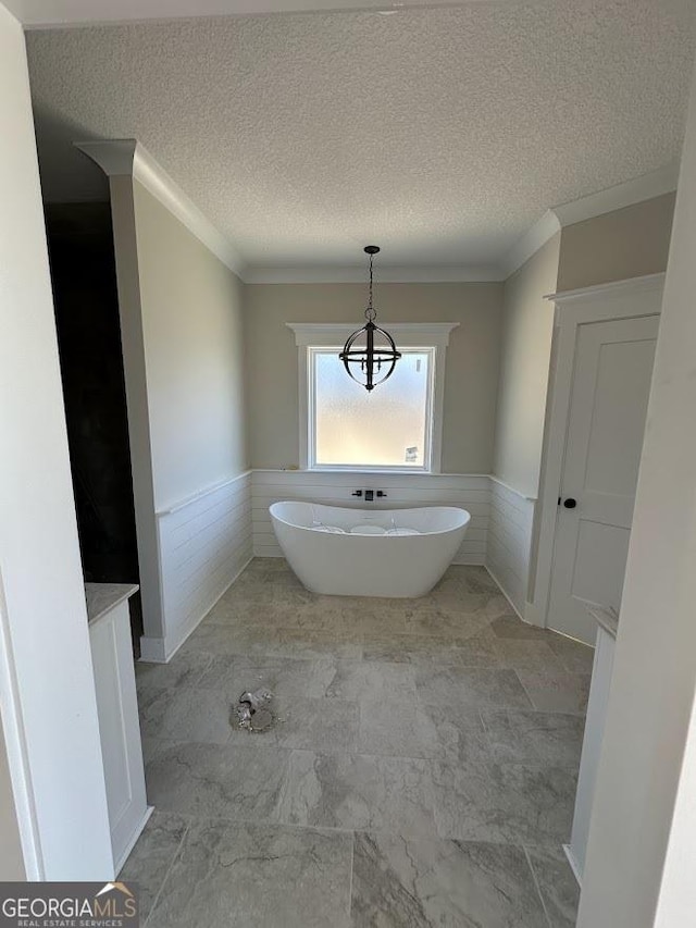 full bath with a textured ceiling, a soaking tub, crown molding, and wainscoting