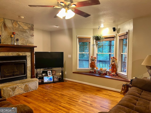 living room featuring a stone fireplace, light hardwood / wood-style floors, a textured ceiling, and ceiling fan