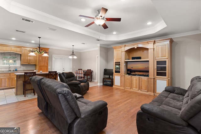 living room featuring sink, light wood-type flooring, ceiling fan with notable chandelier, a tray ceiling, and ornamental molding