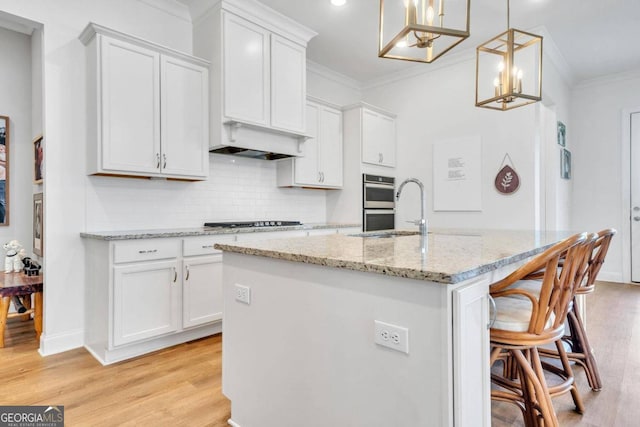 kitchen with white cabinetry, light hardwood / wood-style floors, decorative light fixtures, crown molding, and a center island with sink