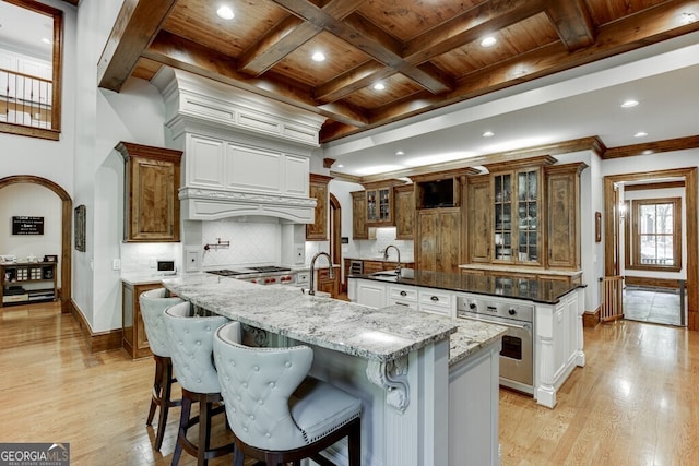 kitchen with tasteful backsplash, white cabinets, a center island with sink, wooden ceiling, and stainless steel oven