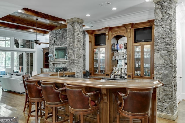 bar with a stone fireplace, ornate columns, light hardwood / wood-style flooring, ornamental molding, and ceiling fan