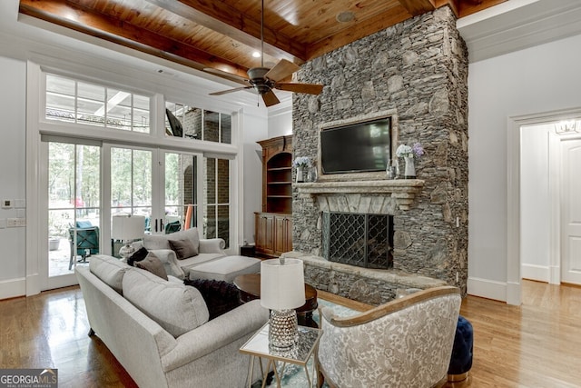 living room with wood ceiling, light hardwood / wood-style flooring, ceiling fan, a stone fireplace, and beamed ceiling