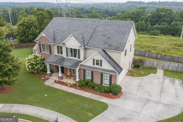 view of front of property featuring brick siding, roof with shingles, covered porch, a front yard, and fence