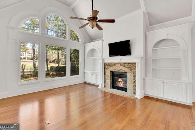 unfurnished living room featuring a stone fireplace, light wood-type flooring, built in shelves, high vaulted ceiling, and ceiling fan