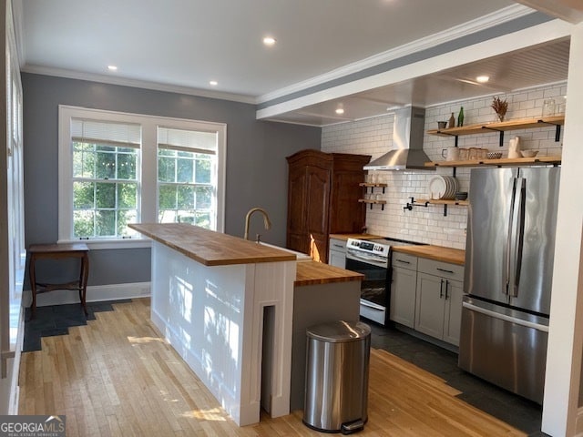 kitchen with wall chimney range hood, an island with sink, wood counters, light hardwood / wood-style flooring, and stainless steel appliances