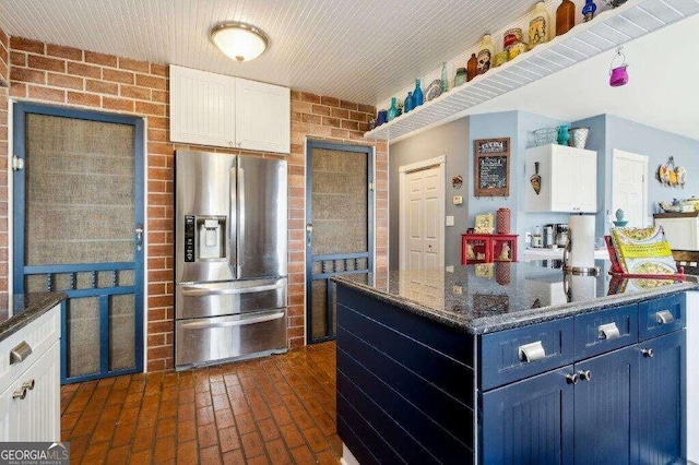 kitchen featuring white cabinetry, blue cabinetry, dark stone countertops, stainless steel refrigerator with ice dispenser, and brick wall