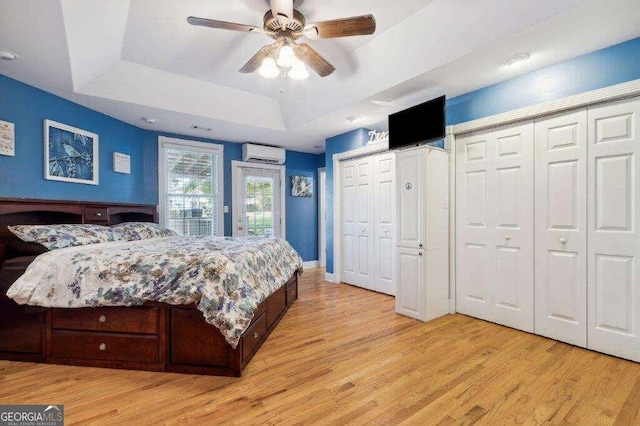 bedroom with light wood-type flooring, a raised ceiling, multiple closets, ceiling fan, and a wall unit AC