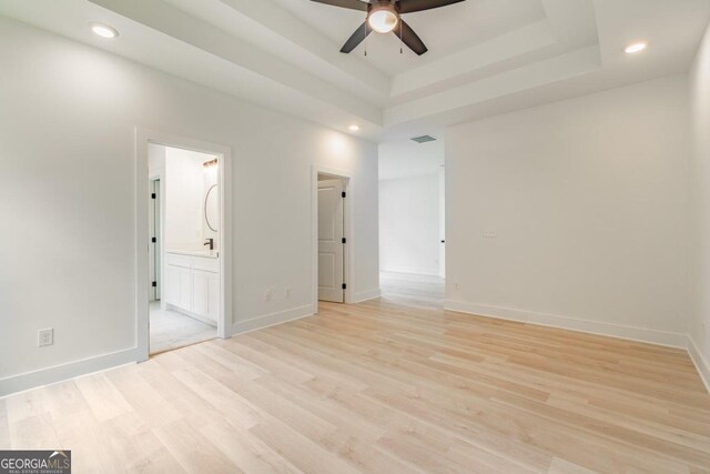 unfurnished bedroom featuring connected bathroom, ceiling fan, a tray ceiling, and light wood-type flooring