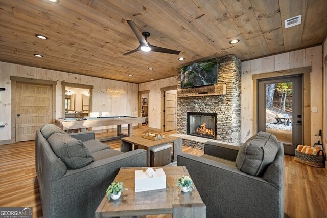 living room featuring wood walls, a stone fireplace, hardwood / wood-style floors, ceiling fan, and wooden ceiling