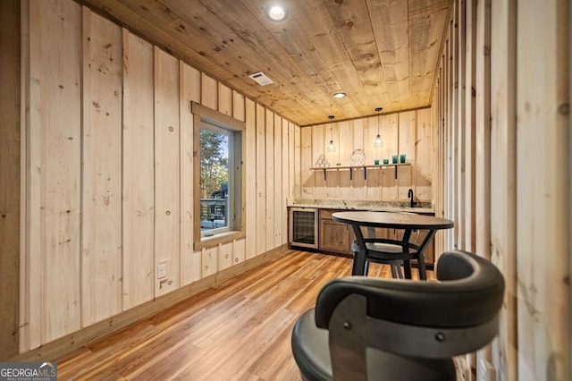 bar with light brown cabinets, wooden walls, wooden ceiling, beverage cooler, and light hardwood / wood-style floors