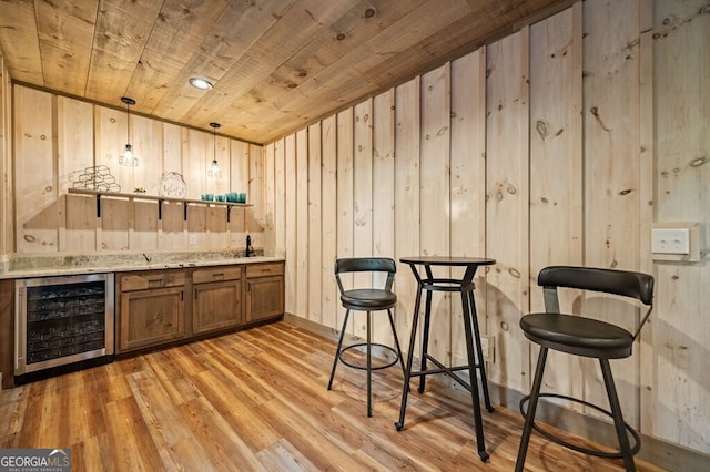 bar with wood walls, light wood-type flooring, wooden ceiling, decorative light fixtures, and beverage cooler