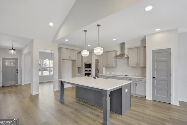 kitchen featuring a kitchen island with sink, wall chimney range hood, gray cabinets, decorative light fixtures, and stainless steel appliances