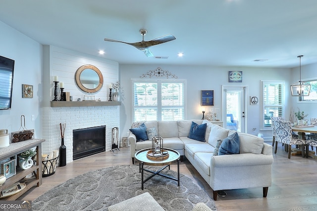 living room featuring light hardwood / wood-style floors, ceiling fan with notable chandelier, and a brick fireplace